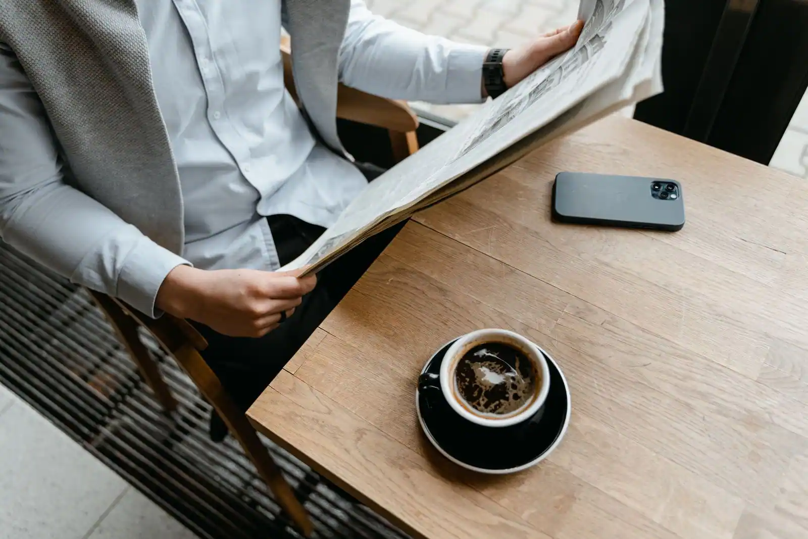 Personne lisant un journal papier à une table, smartphone et tasse de café posés à côté.