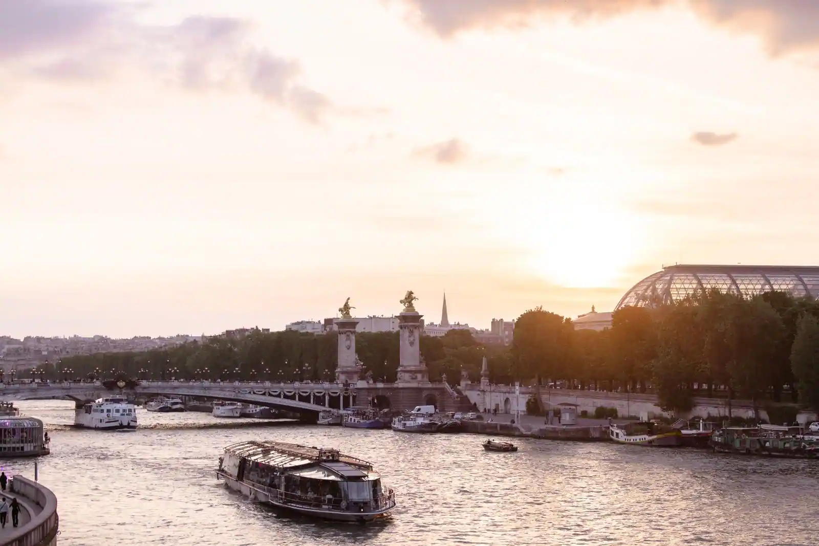 Bateau-promenade vitré naviguant sur la Seine à Paris au coucher du soleil, près d'un pont monumental.