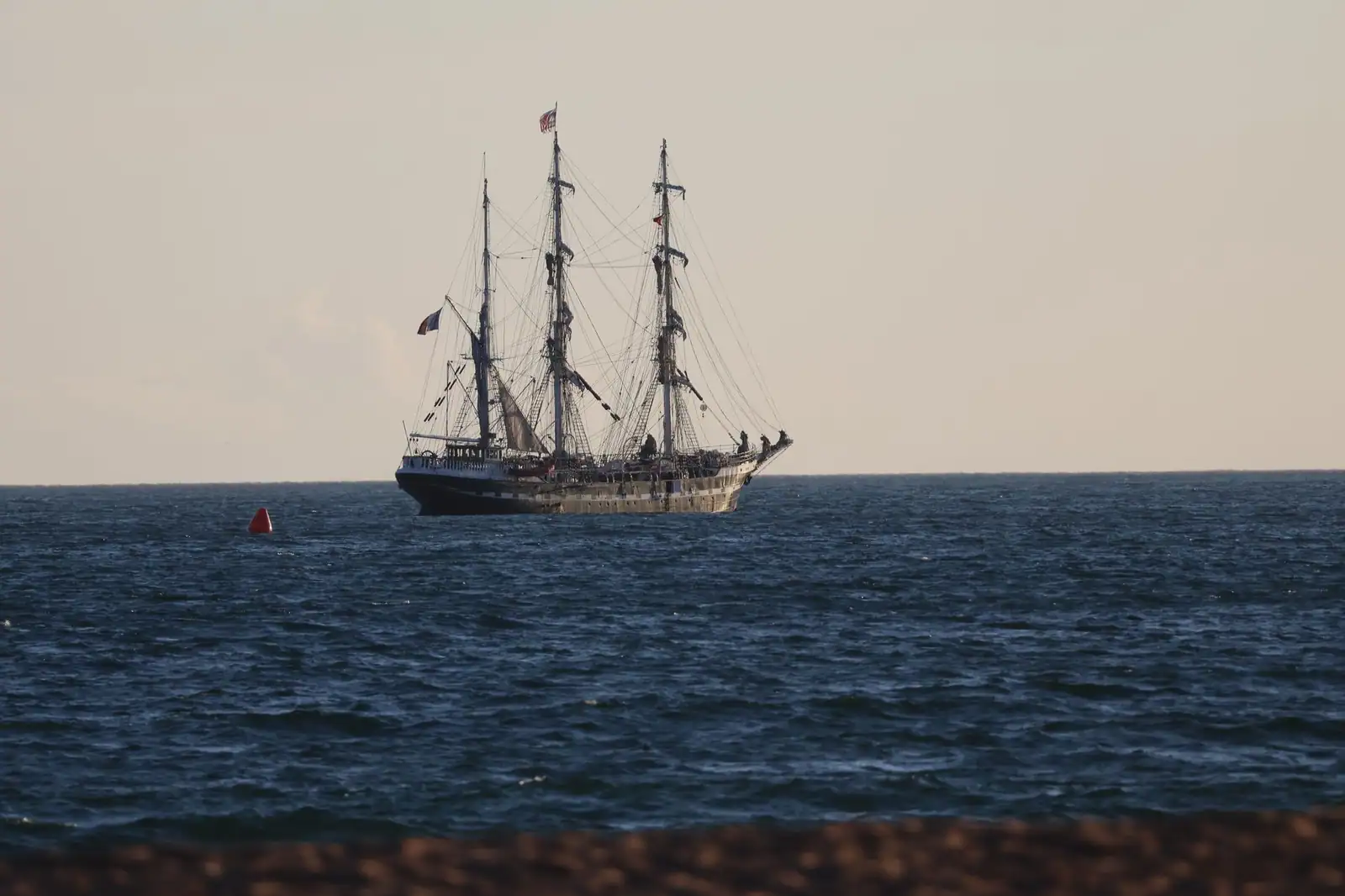 Ancien trois-mâts en bois naviguant au large, mâts et gréement dressés sous un ciel de fin de jour.