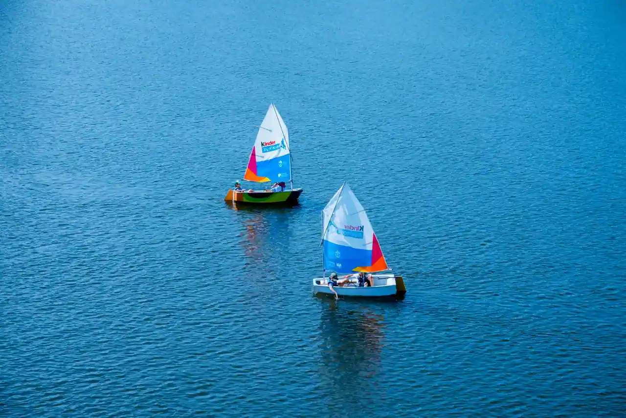 Deux petits dériveurs d'initiation à voile unique, menés par des enfants sur un plan d'eau calme.