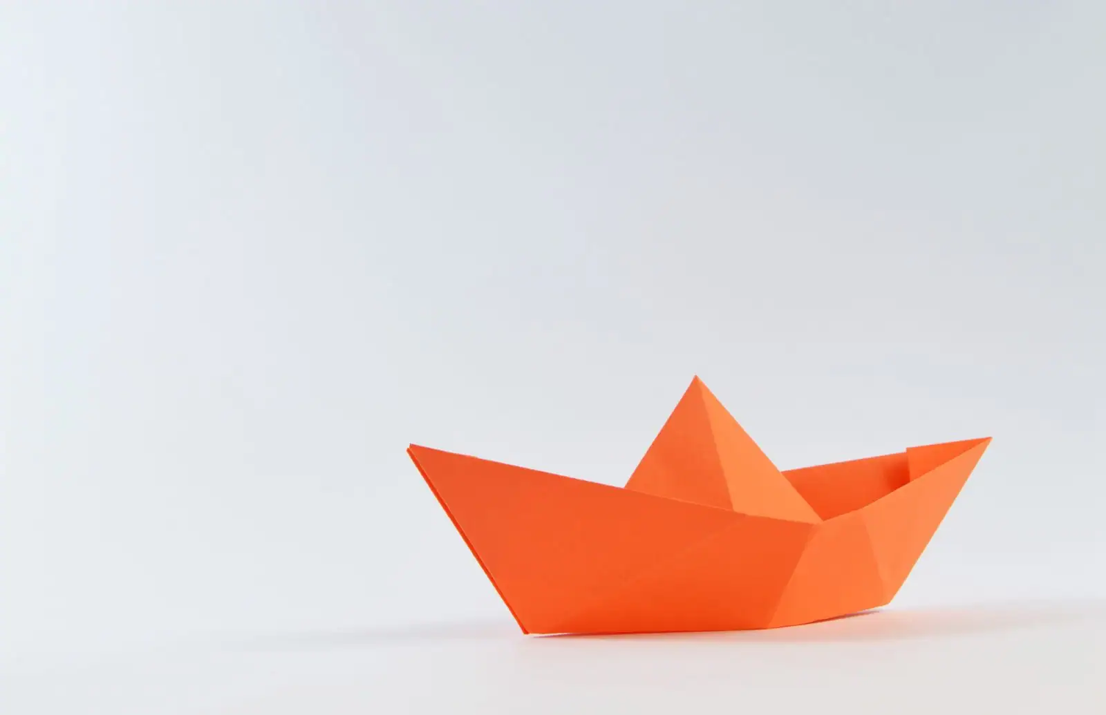 Bateau en papier orange plié selon le modèle classique, posé sur un fond clair épuré.