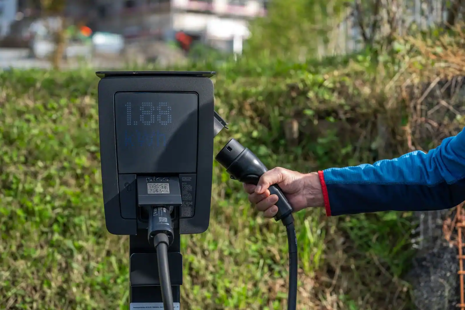 Une main tient le connecteur d'une borne de recharge électrique dont l'écran affiche des kilowattheures