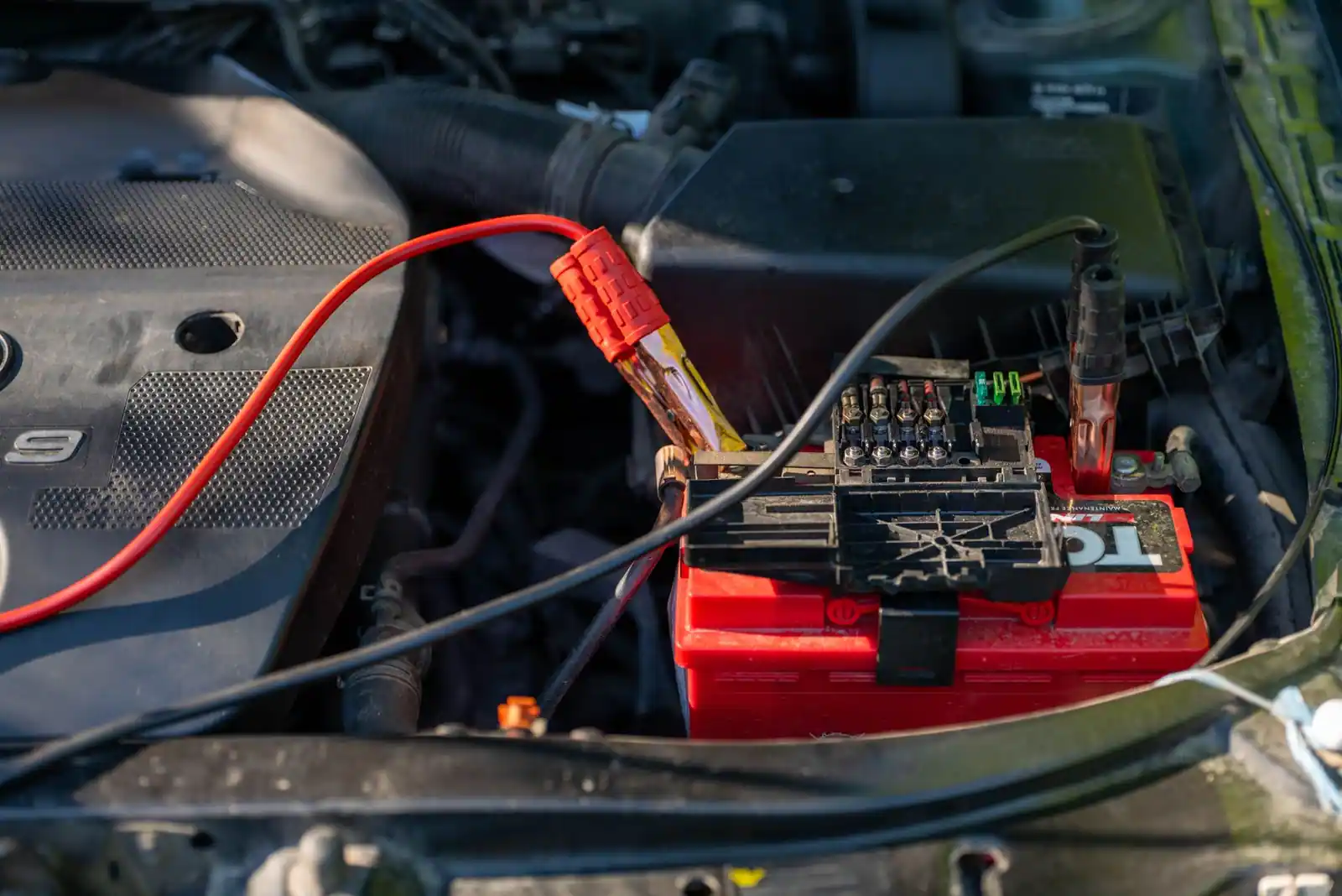 Batterie rouge d'une voiture dans le compartiment moteur, avec câbles et fusibles électriques visibles.