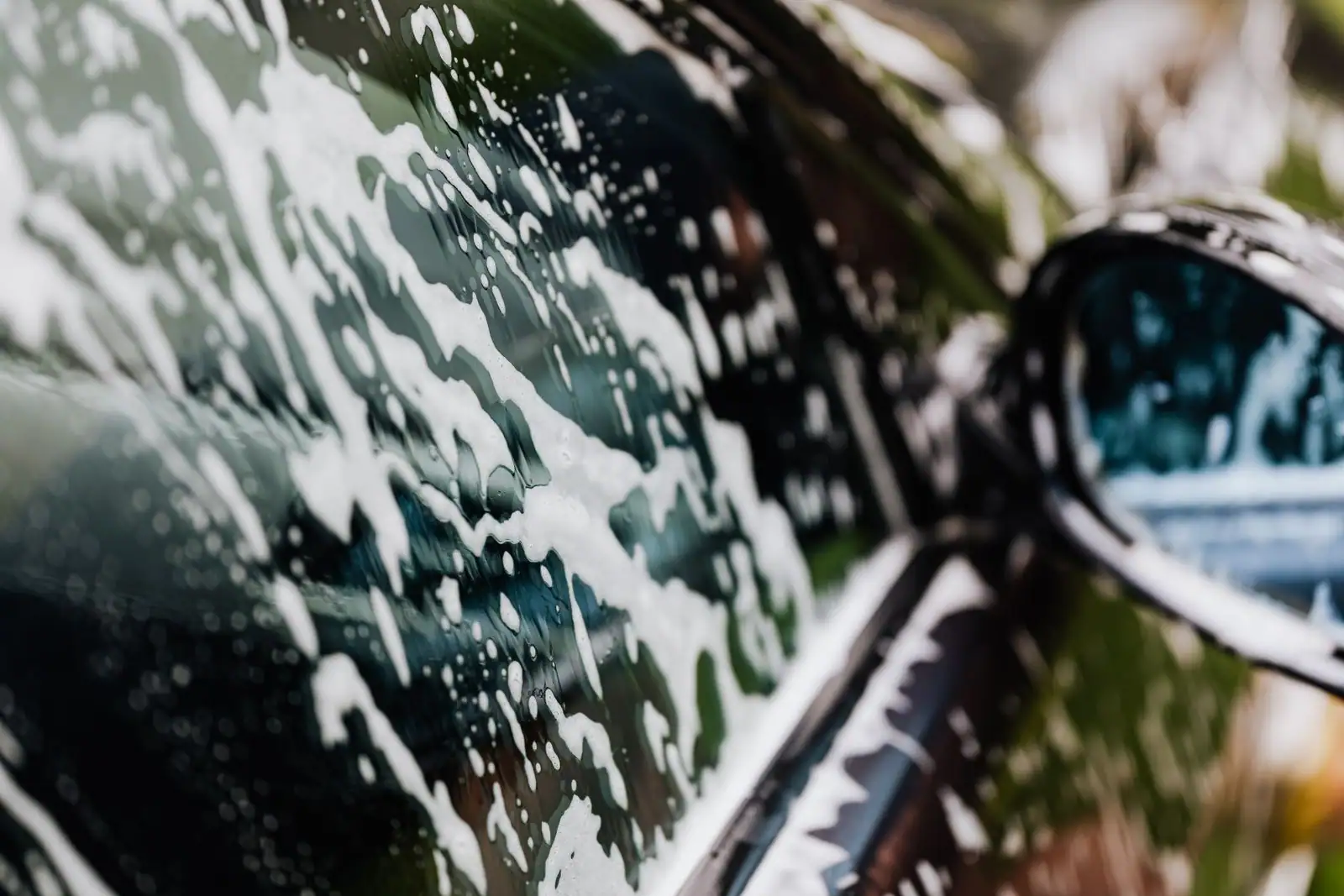 Carrosserie d'une voiture noire couverte de mousse de lavage, vue rapprochée sur la vitre et le rétroviseur.
