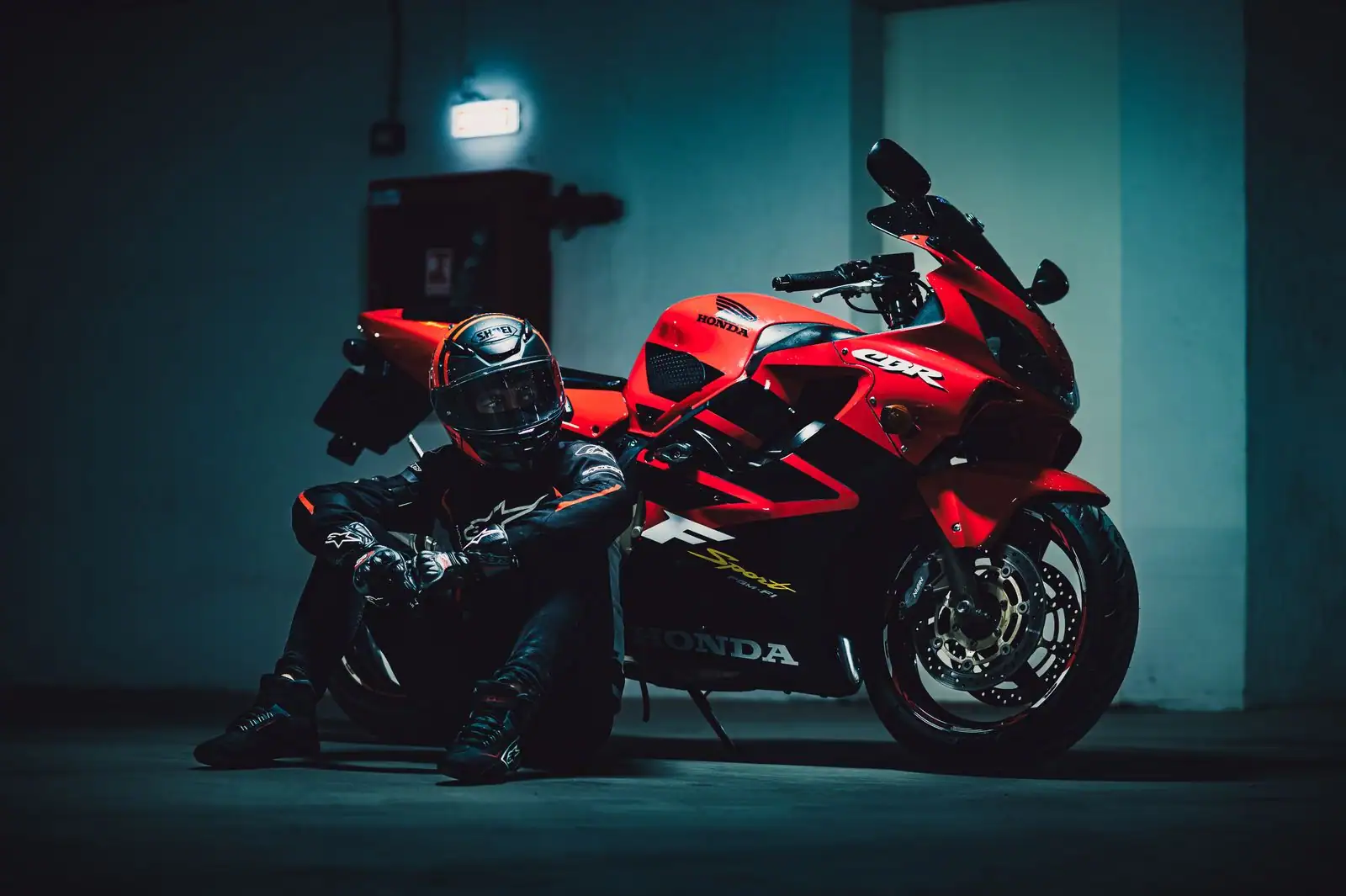 Pilote en combinaison de cuir et casque assis au sol près d'une moto sportive rouge dans un garage