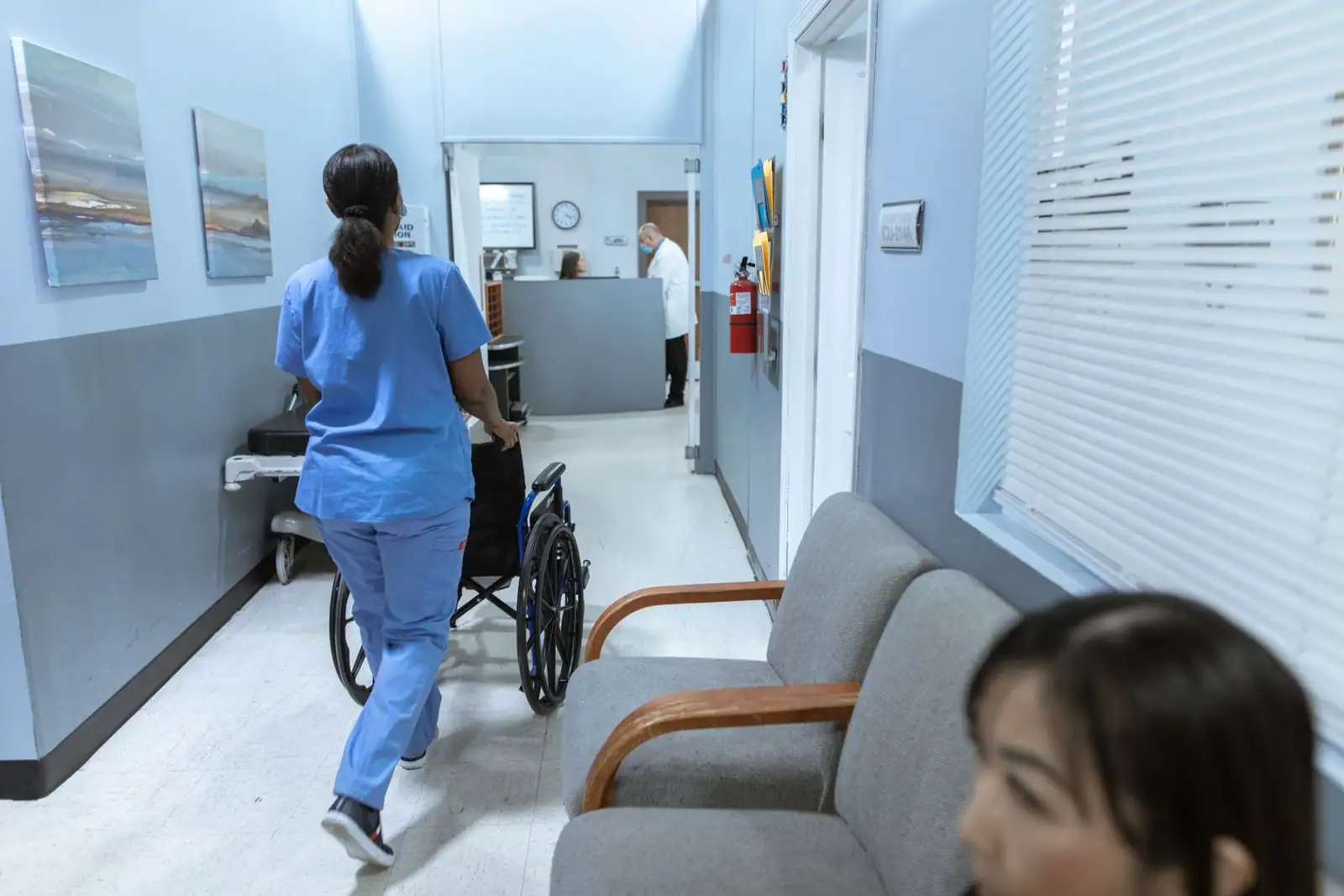 Soignante en tenue bleue poussant un fauteuil roulant dans le couloir d'un établissement de santé.