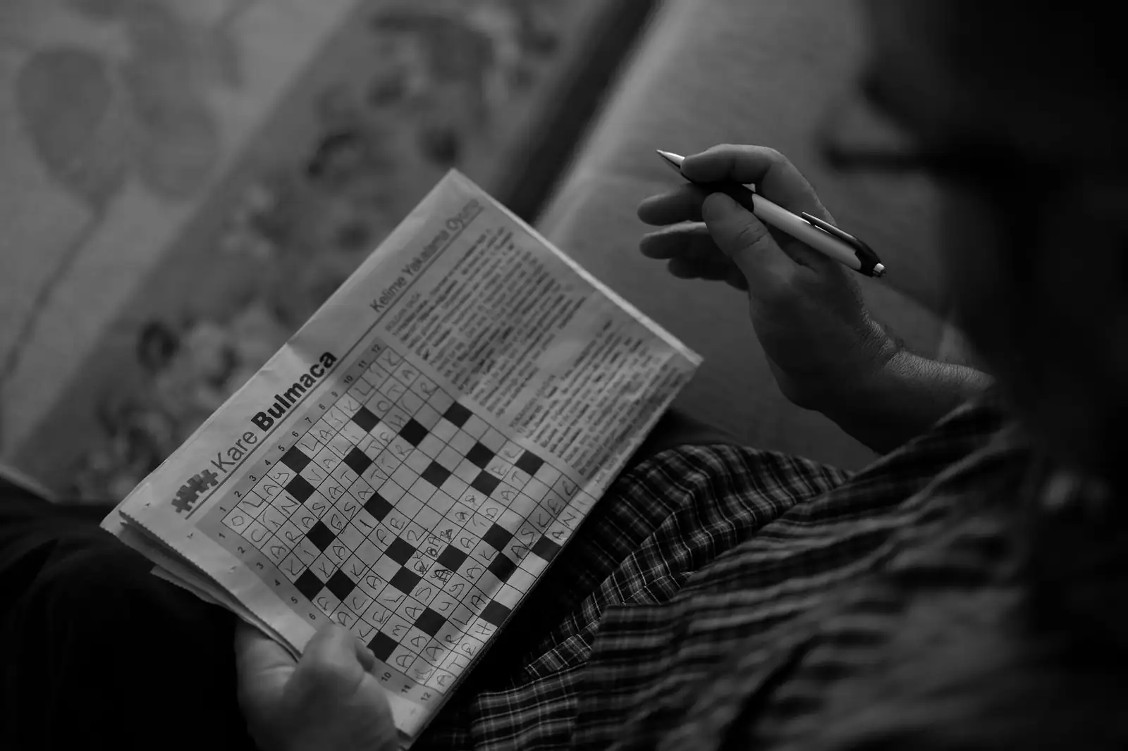 Personne remplissant une grille de mots croisés au stylo, photographie en noir et blanc.