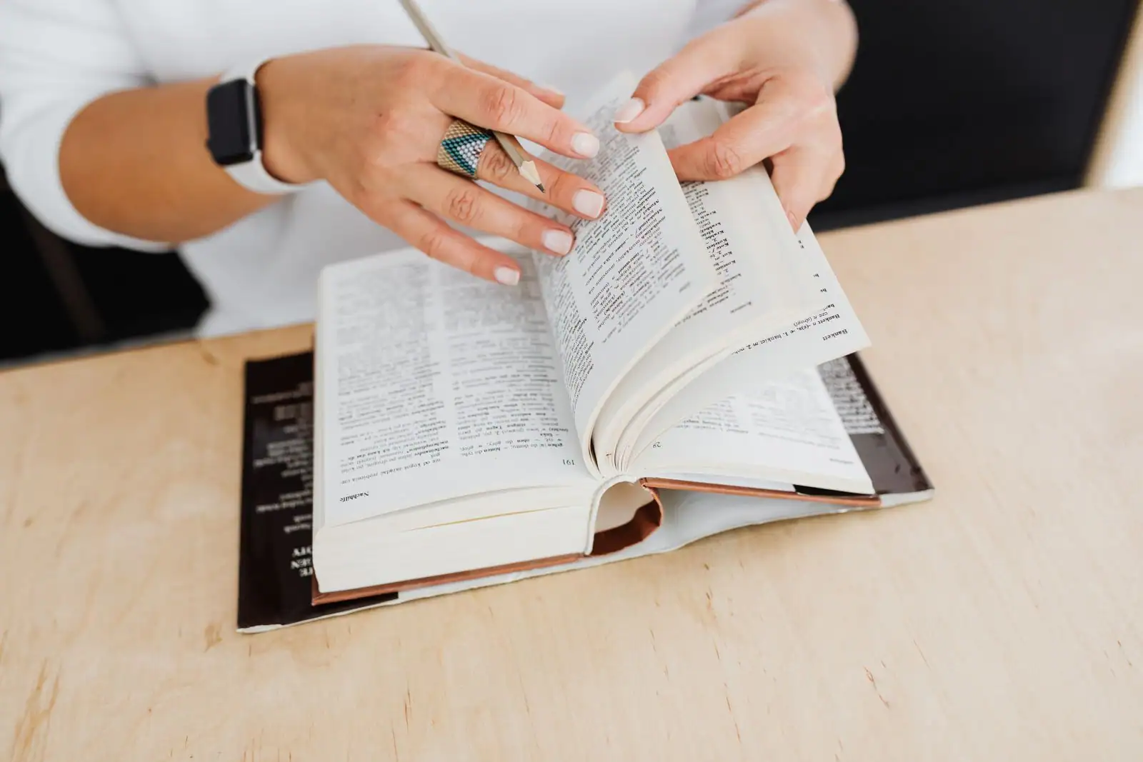 Mains feuilletant un dictionnaire ouvert sur un bureau, un crayon à la main, pour chercher la traduction d'un mot.