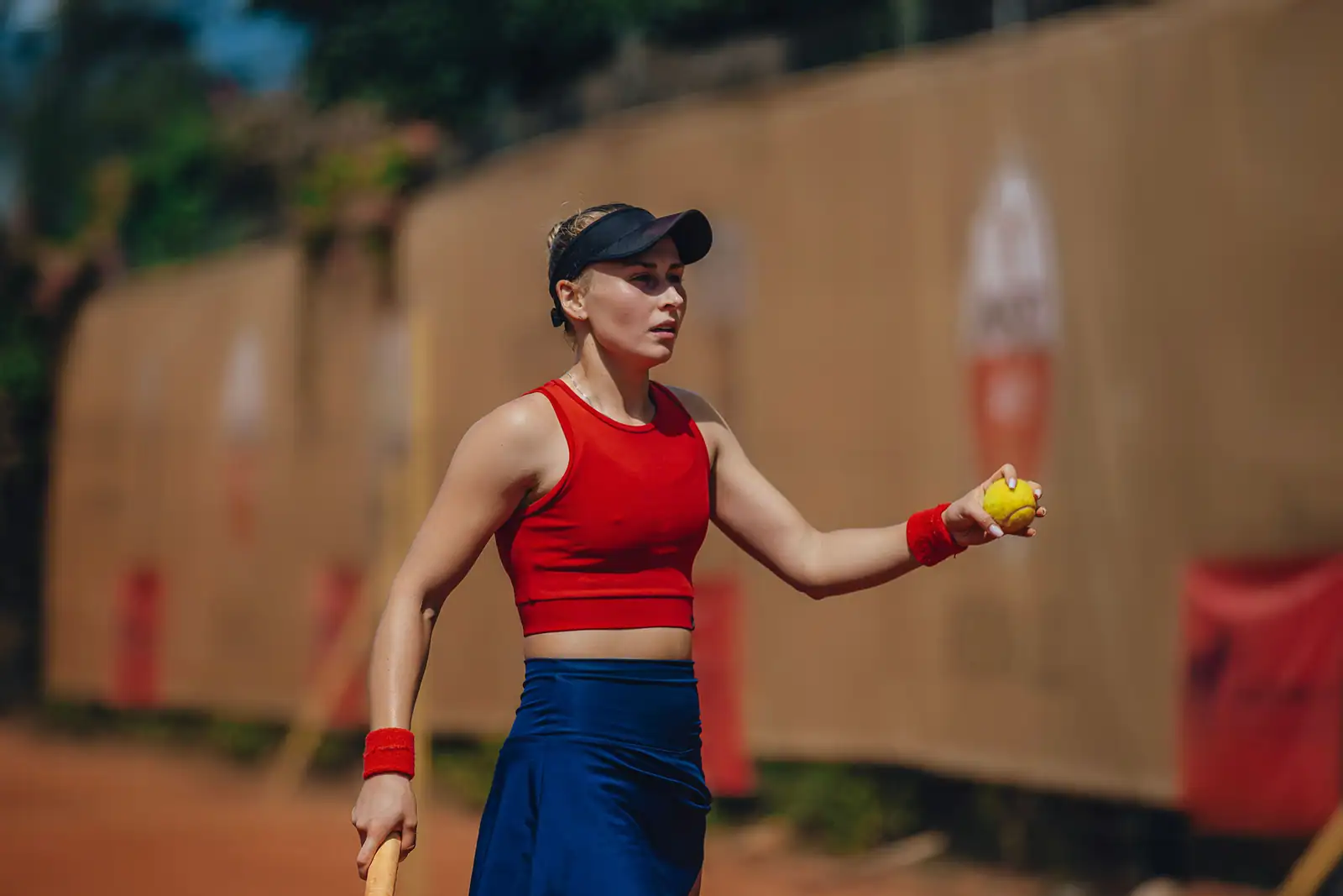Joueuse de tennis balle en main sur un court en terre battue devant les panneaux des sponsors