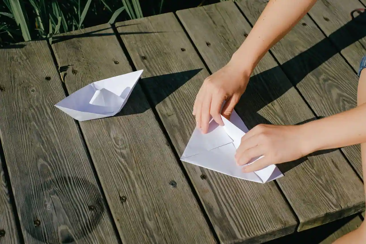 Deux mains plient une feuille blanche sur un ponton en bois, à côté d'un bateau en papier terminé.