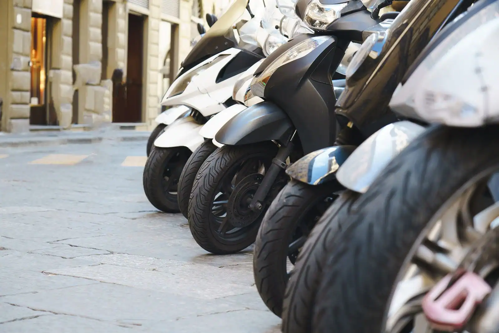 Rangée de scooters urbains garés en épi le long d'une rue pavée, roues avant et antivol au premier plan.