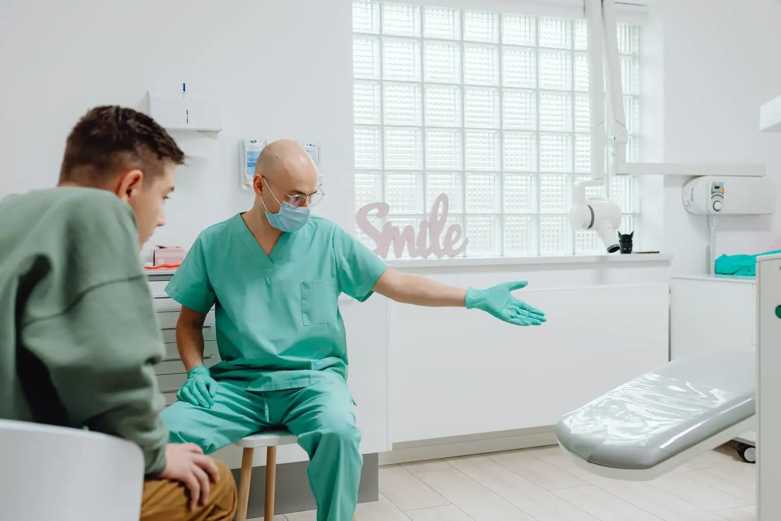 Un praticien en tenue de cabinet dentaire invite un jeune patient à s'installer dans le fauteuil