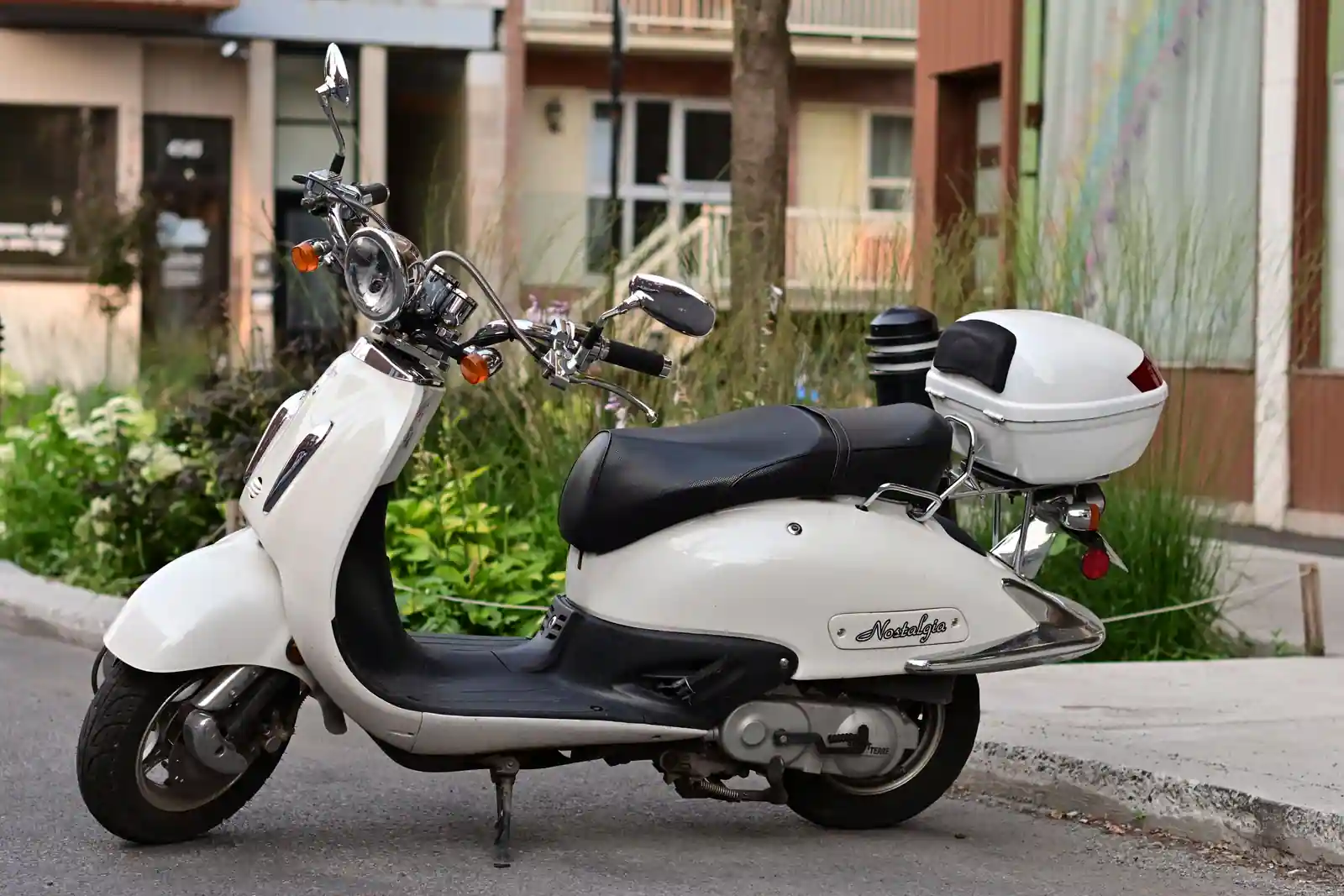 Un scooter urbain blanc garé le long d'un trottoir, top-case monté et carter de transmission apparent.