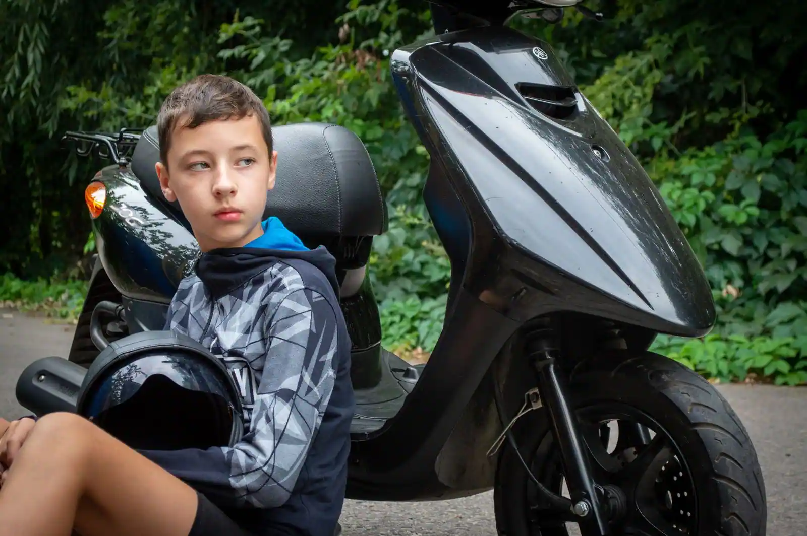 Adolescent assis au sol, casque à la main, près d'un scooter 50 noir à carénage anguleux.