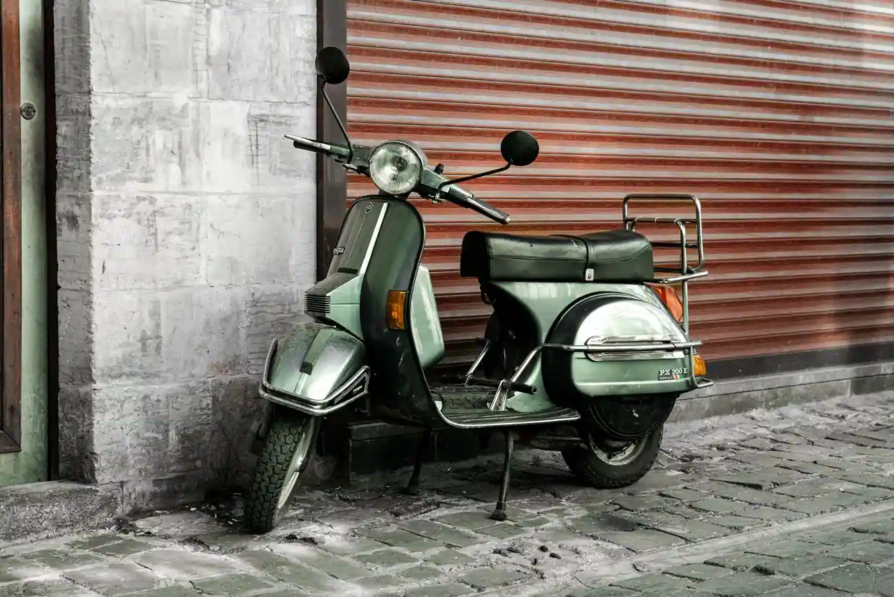 Scooter d'époque vert garé contre un rideau métallique baissé, dans une ruelle pavée