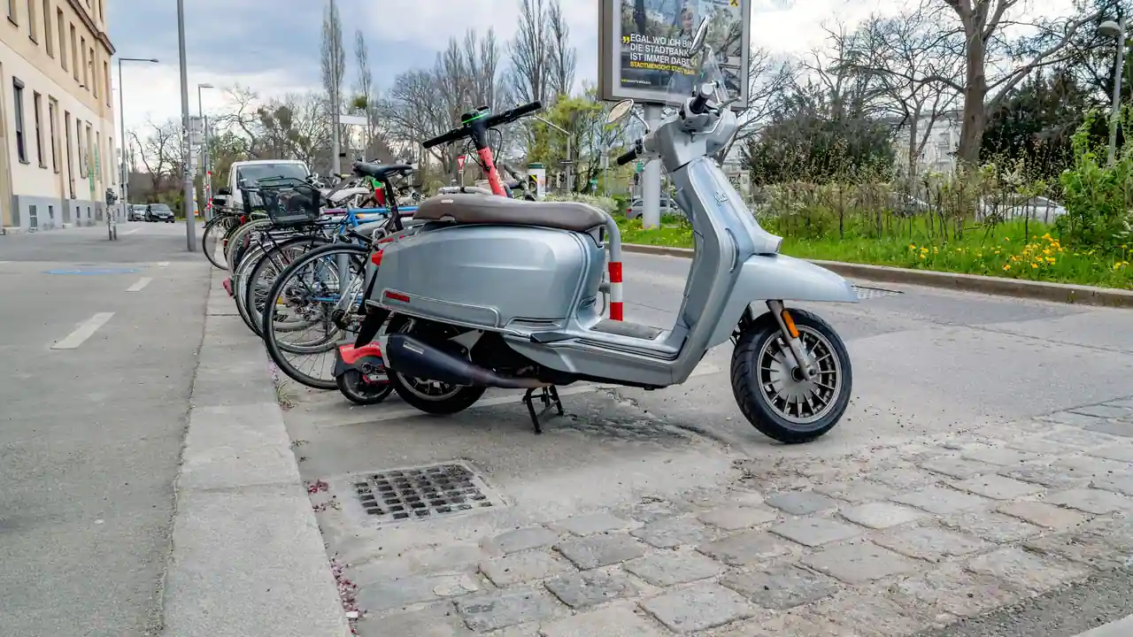 Scooter gris garé le long d'un trottoir, roue avant posée sur les pavés du caniveau, vélos rangés derrière.