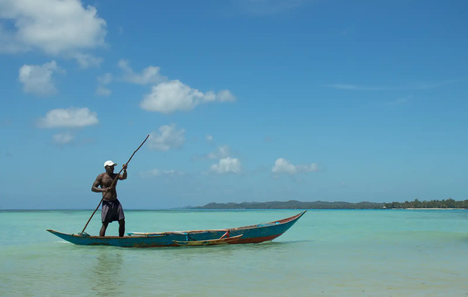 Un pêcheur pousse à la perche une pirogue à balancier unique sur un lagon peu profond.