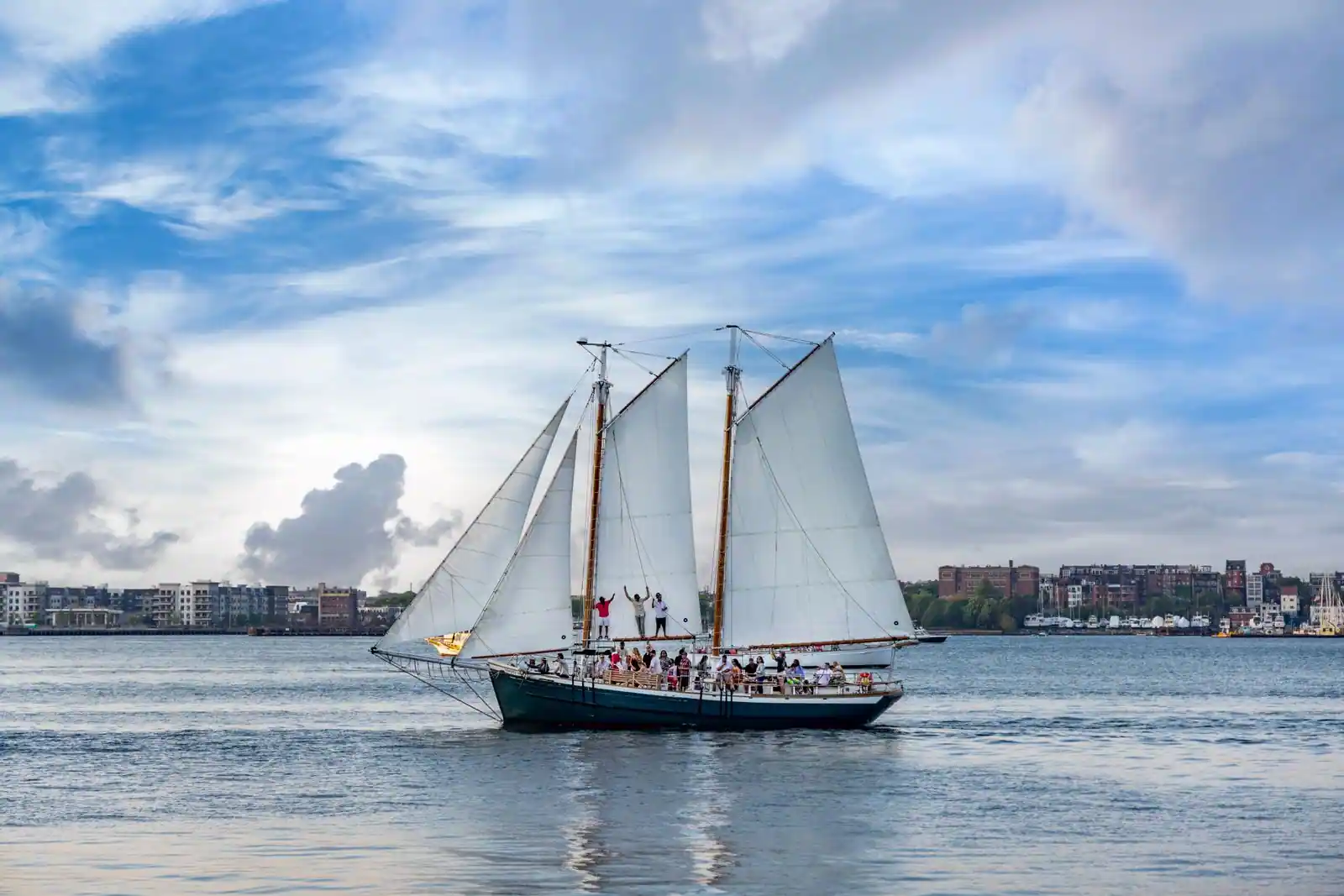Goélette à deux mâts toutes voiles dehors devant un front de mer urbain, passagers debout sur le pont
